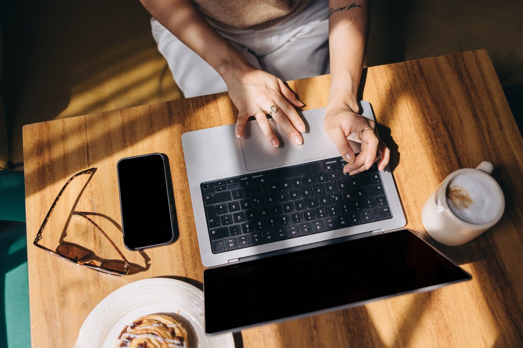 Freelancer working remotely on a laptop at a sunny café table with coffee, sunglasses, and pastry — symbol of flexible work and digital nomad lifestyle.
