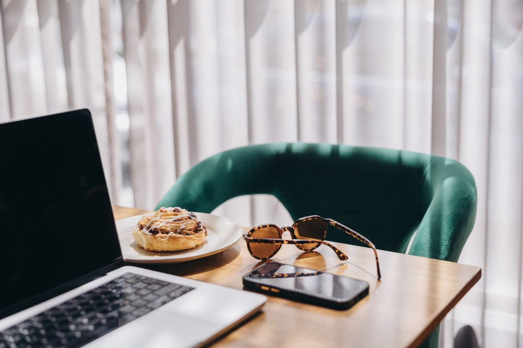 Laptop on a table beside a velvet chair and a cinnamon roll — stylish workation setup.
